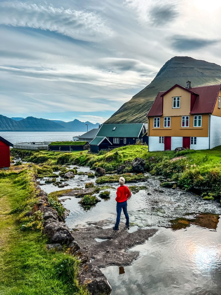 kobiece podróże solo, kobieta w czerwonym swetrze stoi w strumieni, na horyzoncie wysokie góry wyrastające z morza, fiordy, drewniane kolorowe domki 